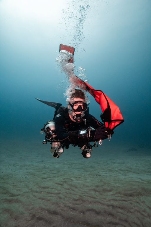 Underwater, a focused dive instructor in a sidemount configuration inflates his DSMB 